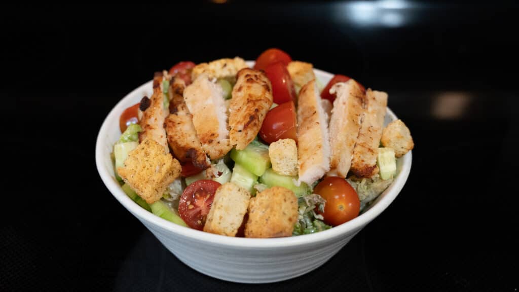 Rotisserie Chicken Caesar Salad in a white stonewash bowl with cucumbers and tomatoes