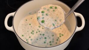 Ladle lifting creamy chicken gravy with peas from a cream enameled cast iron Dutch oven