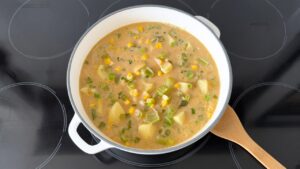 Creamy corn chowder simmering in a white enameled Dutch oven with a wooden spoon