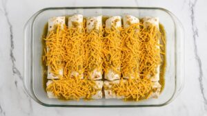 Eight rolled beef enchiladas topped with green chili sauce and shredded cheddar in a 9x13 glass baking dish, ready for the oven