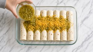 Pouring green chili sauce over rolled chicken enchiladas in a 9x13 glass baking dish