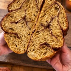 Halved pumpkin cinnamon swirl sourdough loaf showing the swirled crumb on a rustic wood table with a pumpkin, white vase and candle
